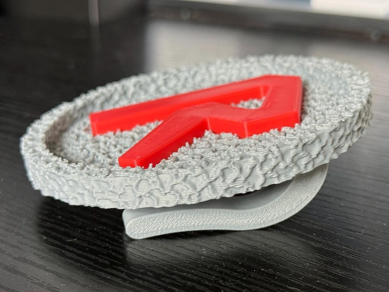 May include: A gray, textured coaster with a raised red letter "R" in the center. The coaster has a unique, patterned surface and a curved, gray base. The design is modern and minimalist.