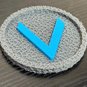 Puede incluir: Un posavasos circular gris con un patr&oacute;n texturizado en forma de red. Una forma angular azul brillante, que se asemeja a la letra "V", est&aacute; centrada en el posavasos. El posavasos probablemente est&aacute; hecho de un material impreso en 3D.