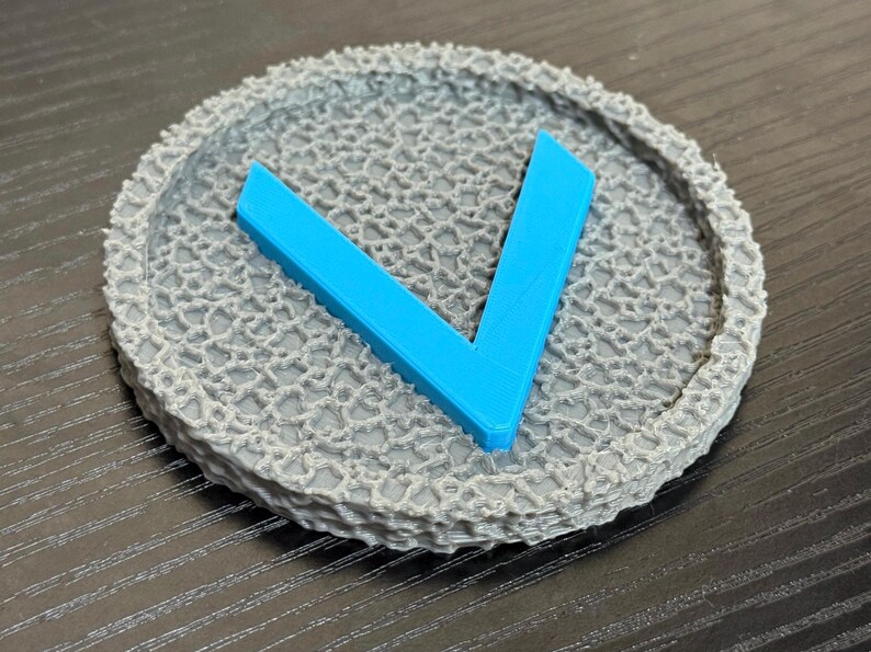Puede incluir: Un posavasos circular gris con un patr&oacute;n texturizado en forma de red. Una forma angular azul brillante, que se asemeja a la letra "V", est&aacute; centrada en el posavasos. El posavasos probablemente est&aacute; hecho de un material impreso en 3D.