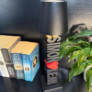 May include: A black lamp with a black lampshade sits on a wooden base with the names "Simon" and "Leah" spelled out in black letters. A red heart is between the names. The lamp is on a table with a stack of books.