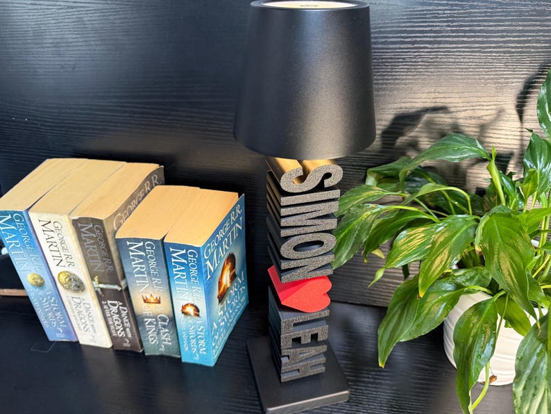 May include: A black lamp with a black lampshade sits on a wooden base with the names "Simon" and "Leah" spelled out in black letters. A red heart is between the names. The lamp is on a table with a stack of books.
