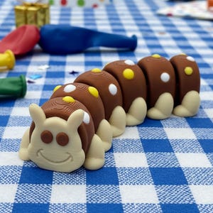 May include: A whimsical, brown and tan caterpillar-shaped figurine with white and yellow accents. The caterpillar has a smiling face and is set against a blue and white checkered background. Balloons and wrapped gifts are also visible.