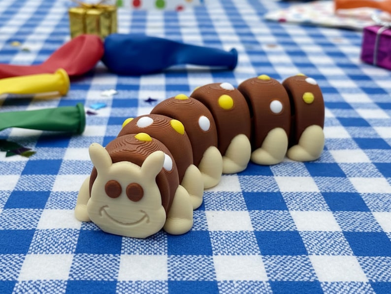 May include: A whimsical, brown and tan caterpillar-shaped figurine with white and yellow accents. The caterpillar has a smiling face and is set against a blue and white checkered background. Balloons and wrapped gifts are also visible.