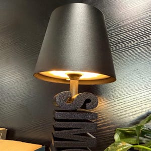 May include: A black lamp with a black fabric shade illuminates a black, stacked, three-dimensional word sculpture that reads "SWIM".