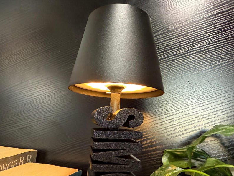 May include: A black lamp with a black fabric shade illuminates a black, stacked, three-dimensional word sculpture that reads "SWIM".