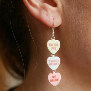May include: A dangling earring featuring three heart-shaped charms. The top charm is pale yellow with the words "CUTIE PIE" in red. The middle charm is white with "SWEET HEART" in red. The bottom charm is pink with "BE MINE" in red.
