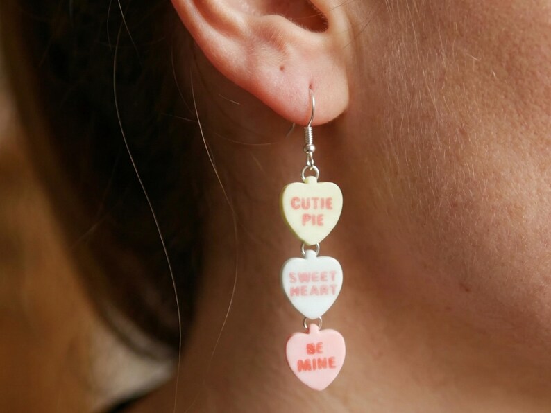 May include: A dangling earring featuring three heart-shaped charms. The top charm is pale yellow with the words "CUTIE PIE" in red. The middle charm is white with "SWEET HEART" in red. The bottom charm is pink with "BE MINE" in red.