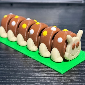 May include: A 3D-printed caterpillar toy with a brown body, white and yellow spots, and off-white legs. The caterpillar rests on a bright green rectangular base. The head has a smiling face and small ears.