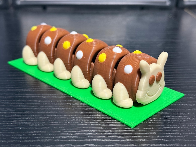 May include: A 3D-printed caterpillar toy with a brown body, white and yellow spots, and off-white legs. The caterpillar rests on a bright green rectangular base. The head has a smiling face and small ears.