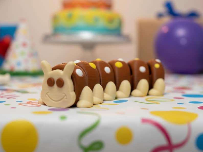 May include: A whimsical, brown caterpillar-shaped cake decoration with white and yellow accents. The smiling caterpillar has a cream-colored face, feet, and antennae. The background includes a birthday cake, party hat, and balloon.