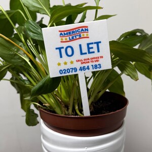 May include: A potted plant with green leaves and a white sign that reads "TO LET." The sign also includes the text "AMERICAN LET'S" and a phone number. The plant is in a brown pot.