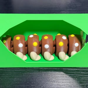 May include: A green, geometric box contains six brown, log-shaped objects with white and yellow circular details. Each log has a beige, rounded base. The objects are arranged neatly inside the box.
