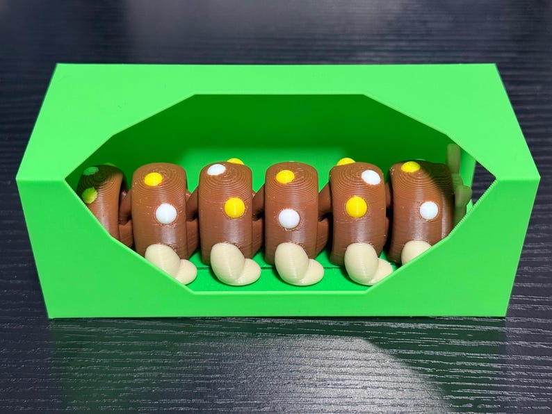 May include: A green, geometric box contains six brown, log-shaped objects with white and yellow circular details. Each log has a beige, rounded base. The objects are arranged neatly inside the box.