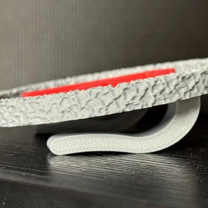 May include: A gray, textured, circular object with a red interior. The object has a curved, gray support. The design has a tire-like pattern around the edge. The object is on a dark surface.