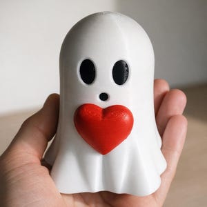 May include: A white ghost figurine with black oval eyes and a small black mouth. A red heart is centered on the ghost's chest. The figurine is held in a hand.