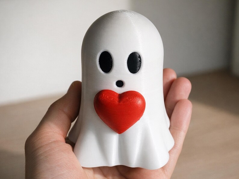 May include: A white ghost figurine with black oval eyes and a small black mouth. A red heart is centered on the ghost's chest. The figurine is held in a hand.