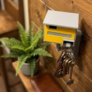 K&ouml;nnte beinhalten: Ein an der Wand befestigter Schl&uuml;sselhalter in Form eines Miniatur-Briefkastens. Der Schl&uuml;sselhalter ist grau und gelb, mit einem kleinen Dach und einem Schlitz f&uuml;r Post. Ein Satz Schl&uuml;ssel h&auml;ngt von unten. Der Halter ist an einer Holzwand befestigt.