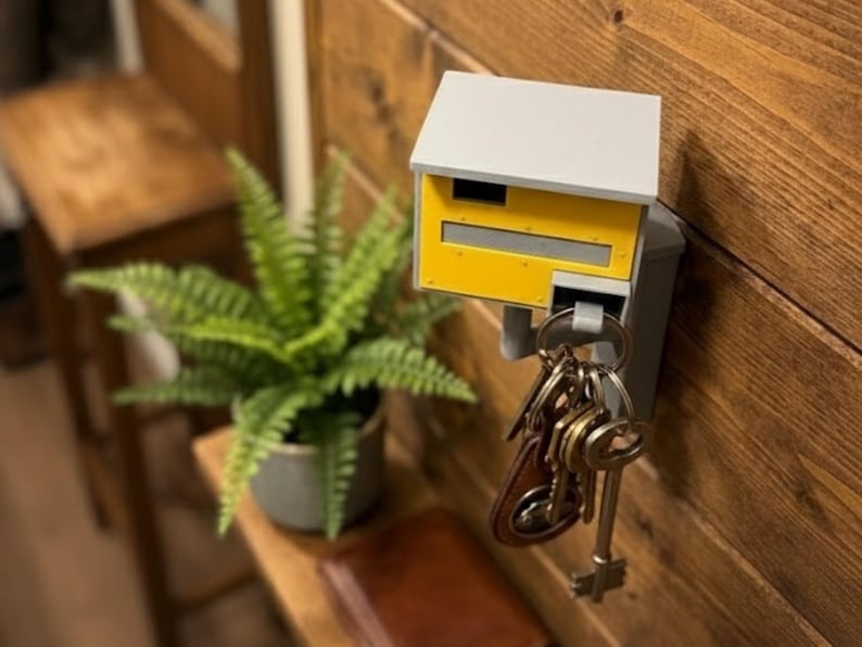 K&ouml;nnte beinhalten: Ein an der Wand befestigter Schl&uuml;sselhalter in Form eines Miniatur-Briefkastens. Der Schl&uuml;sselhalter ist grau und gelb, mit einem kleinen Dach und einem Schlitz f&uuml;r Post. Ein Satz Schl&uuml;ssel h&auml;ngt von unten. Der Halter ist an einer Holzwand befestigt.