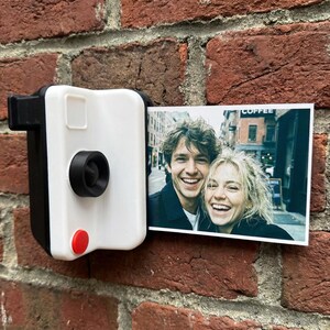 May include: A white and black camera-shaped photo holder with a red button, mounted on a brick wall. A colour photograph of two smiling people is displayed in the holder. The word "COFFEE" is visible in the background.