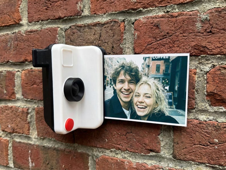 May include: A white and black camera-shaped photo holder with a red button, mounted on a brick wall. A colour photograph of two smiling people is displayed in the holder. The word "COFFEE" is visible in the background.