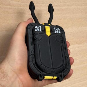 May include: A black and yellow 3D-printed device with two curved, antenna-like protrusions. The device has a rectangular shape with rounded edges and intricate detailing. The design includes yellow accents and white symbols. The device is held in a hand.