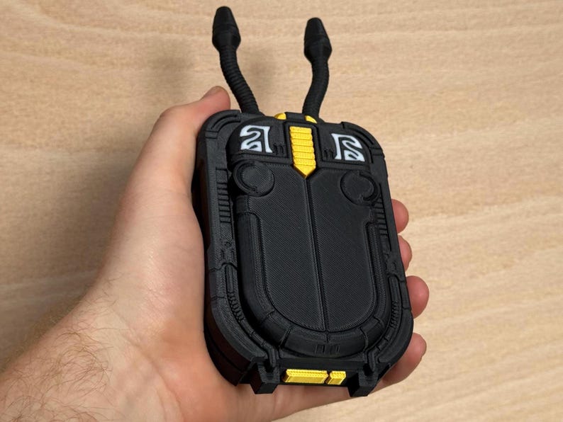 May include: A black and yellow 3D-printed device with two curved, antenna-like protrusions. The device has a rectangular shape with rounded edges and intricate detailing. The design includes yellow accents and white symbols. The device is held in a hand.