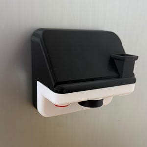 May include: A black and white 3D-printed camera-shaped object with a red button and a black lens. The object is mounted on a metallic surface. The top portion is black, and the bottom is white.