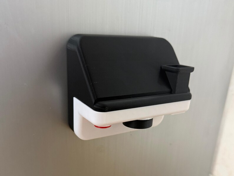 May include: A black and white 3D-printed camera-shaped object with a red button and a black lens. The object is mounted on a metallic surface. The top portion is black, and the bottom is white.