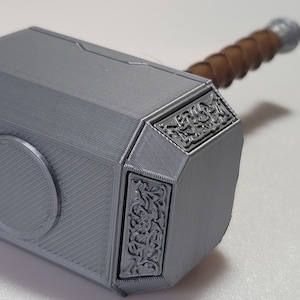 May include: A silver-colored Mjolnir hammer replica with a brown wooden handle. The hammer head has intricate details and a circular design on the front.
