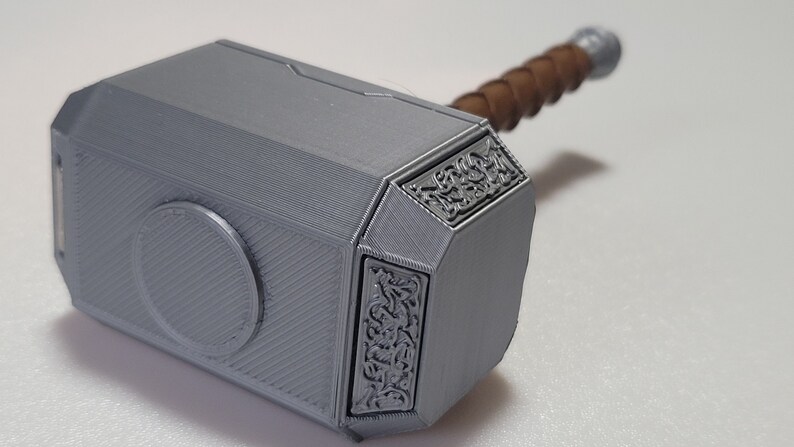 May include: A silver-colored Mjolnir hammer replica with a brown wooden handle. The hammer head has intricate details and a circular design on the front.