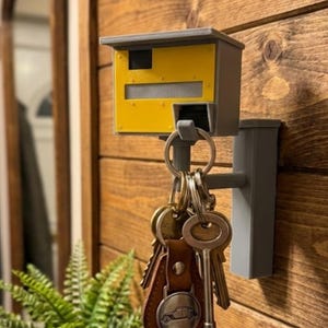 K&ouml;nnte beinhalten: Ein grauer und gelber Schl&uuml;sselhalter in Form eines Blitzers, montiert an einer Holzwand. Ein Satz Schl&uuml;ssel h&auml;ngt an dem Schl&uuml;sselhalter. Der Schl&uuml;sselhalter hat eine rechteckige gelbe Vorderseite mit einem grauen Ober- und Unterteil.