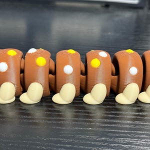 May include: A brown articulated toy with a series of connected cylindrical segments. Each segment has a white and yellow dot, and a beige foot-like shape. The toy is designed to bend and move.