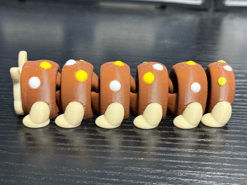 May include: A brown articulated toy with a series of connected cylindrical segments. Each segment has a white and yellow dot, and a beige foot-like shape. The toy is designed to bend and move.