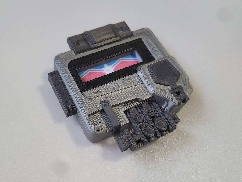 May include: A gray 3D printed device with a black screen and a small flag displayed on the screen. The device has several buttons and a textured surface.
