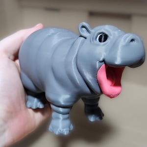 May include: A 3D printed gray hippopotamus figurine with an open mouth and a pink tongue. The hippopotamus has a cartoonish appearance with large eyes.