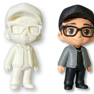 May include: Two figurines of a person wearing glasses and a cap. One is unpainted, the other is painted with a black cap, black glasses, a gray jacket, black pants, and a black shirt. The figurines are standing.