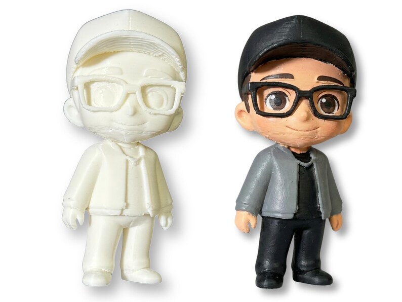 May include: Two figurines of a person wearing glasses and a cap. One is unpainted, the other is painted with a black cap, black glasses, a gray jacket, black pants, and a black shirt. The figurines are standing.