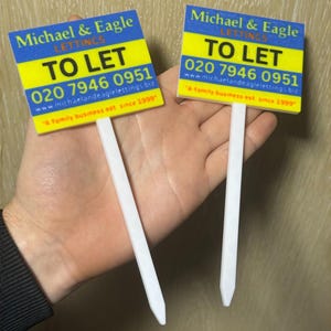 May include: Two "To Let" signs with white stakes. The signs have a blue and yellow background with the text "Michael & Eagle LETTINGS" and a phone number. The signs are for property rentals.
