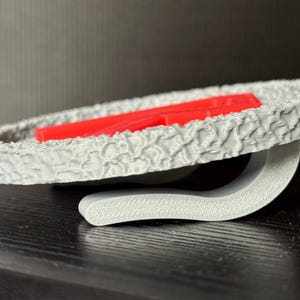 May include: A gray, textured, circular object with a red rectangular piece inside. The object is supported by a curved gray stand. The design has a unique, patterned surface.