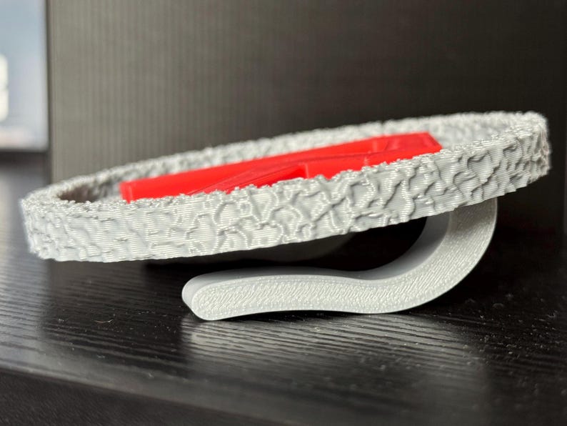May include: A gray, textured, circular object with a red rectangular piece inside. The object is supported by a curved gray stand. The design has a unique, patterned surface.