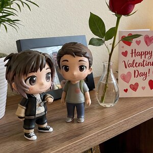 May include: Two anime-style figurines of young people stand on a wooden surface. A Valentine's Day card with hearts and the text "Happy Valentine's Day!" is next to a red rose in a small vase. A framed photo is in the background.