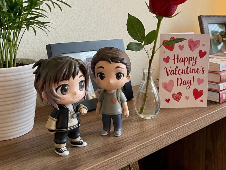 May include: Two anime-style figurines of young people stand on a wooden surface. A Valentine's Day card with hearts and the text "Happy Valentine's Day!" is next to a red rose in a small vase. A framed photo is in the background.