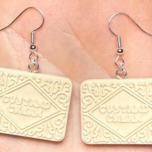 Puede incluir: Un par de pendientes con forma de galletas Custard Cream. Las galletas de color crema tienen forma rectangular con patrones en relieve y la inscripci&oacute;n "Custard Cream". Cuelgan de ganchos plateados.