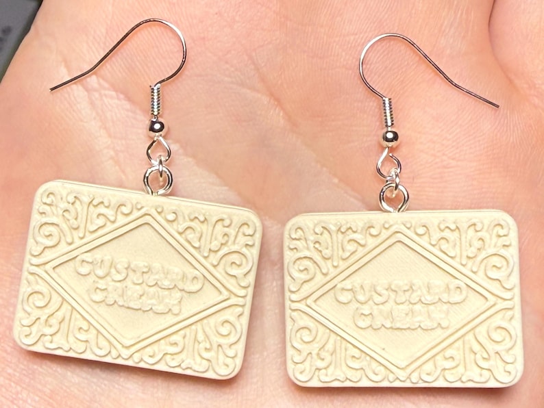 Puede incluir: Un par de pendientes con forma de galletas Custard Cream. Las galletas de color crema tienen forma rectangular con patrones en relieve y la inscripci&oacute;n "Custard Cream". Cuelgan de ganchos plateados.