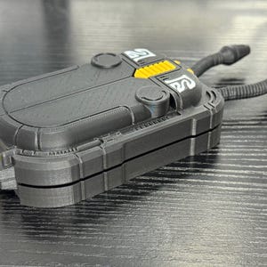May include: A black and yellow rectangular device with two curved, black tubes extending from the top. The device has a textured surface and a hinged design. The top features yellow accents and white symbols.