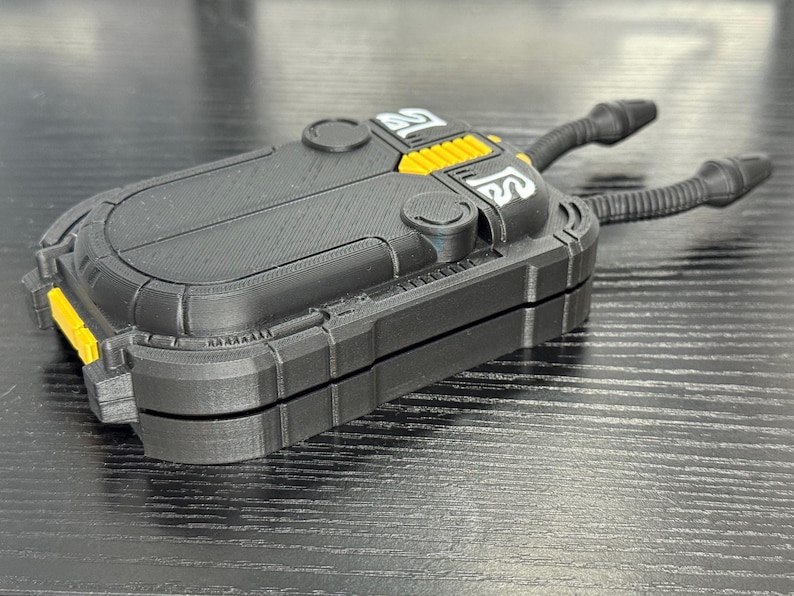 May include: A black and yellow rectangular device with two curved, black tubes extending from the top. The device has a textured surface and a hinged design. The top features yellow accents and white symbols.