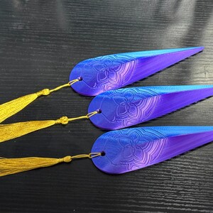 May include: Three blue and purple feather-shaped bookmarks with a floral design and gold tassels. The bookmarks are made of a gradient of colors, transitioning from blue to purple. Each bookmark has a gold tassel attached.