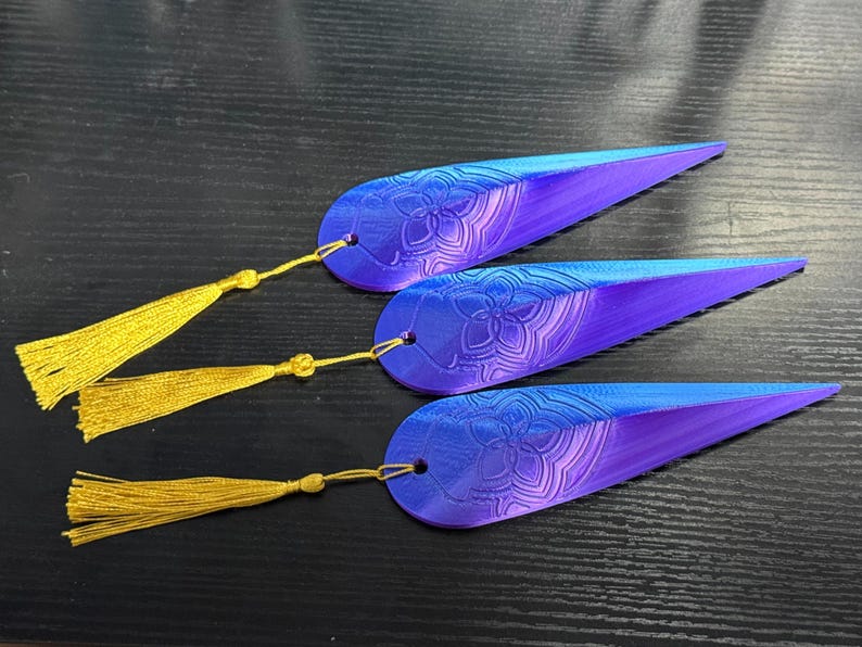 May include: Three blue and purple feather-shaped bookmarks with a floral design and gold tassels. The bookmarks are made of a gradient of colors, transitioning from blue to purple. Each bookmark has a gold tassel attached.