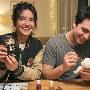 May include: Two people are smiling while painting figurines. One person holds a finished figurine with black, white, and gold accents. The other person is painting a white figurine with a paintbrush and yellow paint.