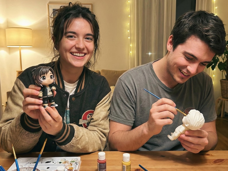 May include: Two people are smiling while painting figurines. One person holds a finished figurine with black, white, and gold accents. The other person is painting a white figurine with a paintbrush and yellow paint.